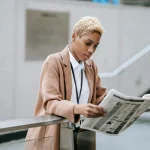 Professional woman reading a newspaper while standing outdoors in an urban setting.