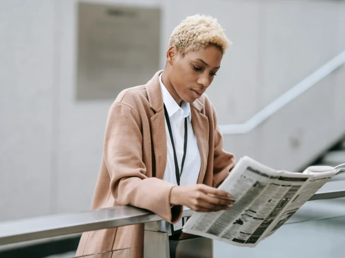 Professional woman reading a newspaper while standing outdoors in an urban setting.